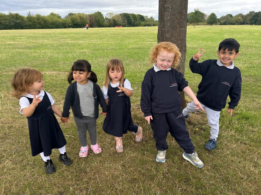 Five children standing together on grass beside a tree.